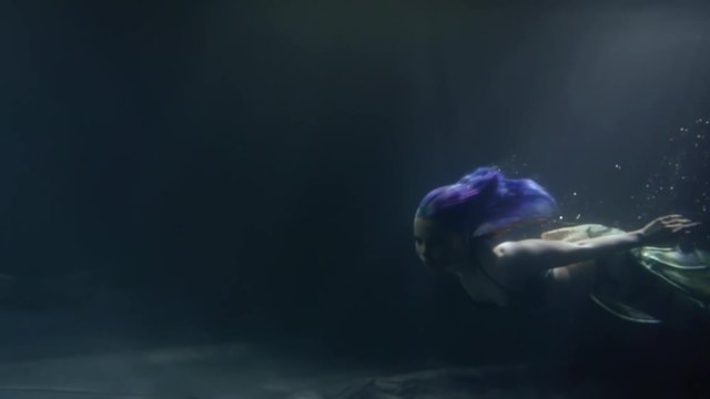 Fantastic shoot of a beautiful model in a mermaid costume swimming by under dim light beams coming throught the water.