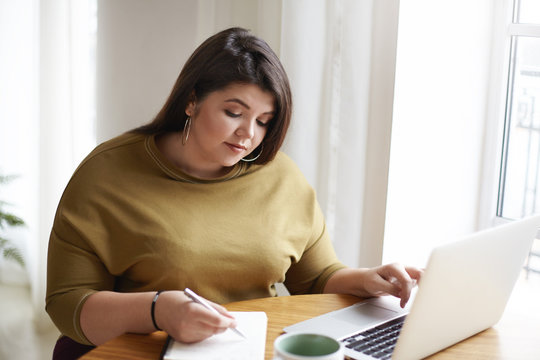Picture of attractive young European female journalist wearng stylish earrings and knitted sweater making notes in diary while working on research for her new article, looking for information online