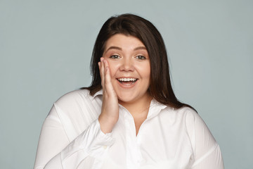 Isolated shot of plus size excited young female employee expressing astonishment and amazement, being unexpectedly promoted, exclaiming, holding hand on cheek. Positive human emotions and feelings