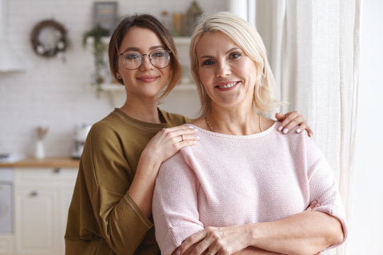 Horizontal image of cute young daughter in stylish hipster eyeglasses embracing her mother with love, spending day together, having happy joyful facial expressions, smiling broadly at camera