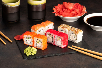 Sushi rolls and sashimi in a black stone plate. Traditional Japanese cuisine with salmon, fish, wasabi, soy sauce, and ginger. Chopsticks and two cups of traditional sake