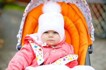 Cute little beautiful baby girl sitting in the pram or stroller on autumn day. Happy healthy child going for a walk on fresh air in warm clothes. Baby with yellow fall maple trees in colorful clothes