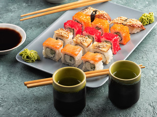 Sushi rolls and sashimi in a grey plate. Traditional Japanese cuisine with salmon, fish, wasabi, soy sauce, and ginger. Chopsticks and two cups of traditional sake