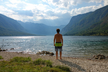 Unrecognizable shirtless man stands on coast and observes the lake and valley.