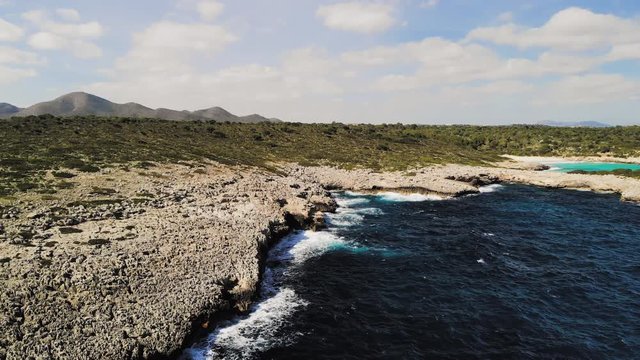 a drone shot flying towards a rocky shore with waves crashing into the rocks and spraying into the air near cala varques in majorca