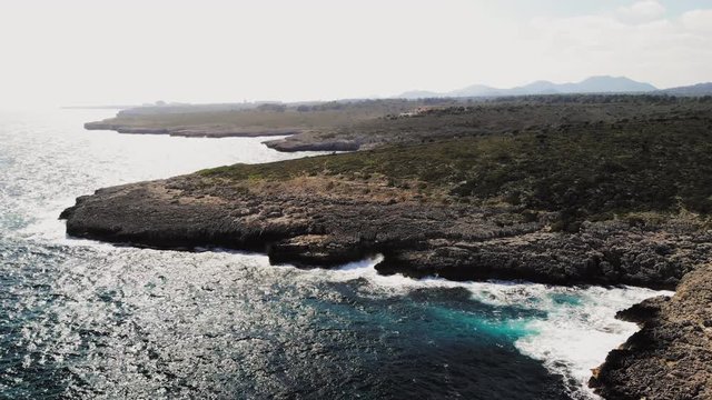 a forward flying drone shot of a large rocky landscape with waves crashing onto the rocks and creating sea spray near cala varques in majorca