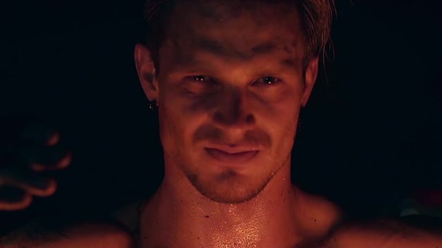 Blond male holds two pois hanging on chains. Male stares at camera. Face and neck are sweaty. Fire reflects on skin. Slow motion shot. Close-up shot.