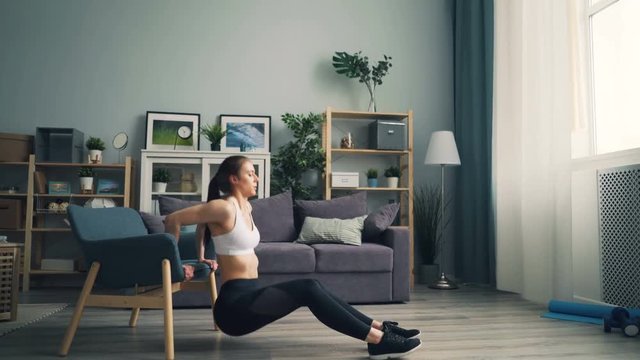 Side view of fit young woman doing pushup dips holding armchair working out in apartment wearing trendy sportswear. People, interior and training concept.