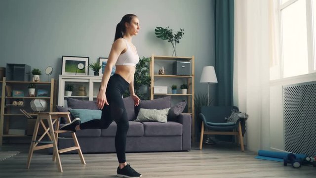 Muscular young woman student is doing squats indoors concentrated on physical exercise training alone. Modern youth, healthy lifestyle and apartment concept.