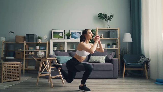 Sporty young lady is squatting on one leg at home exercising alone in studio apartment wearing top, leggings and sneakers. Youth, house and sports concept.