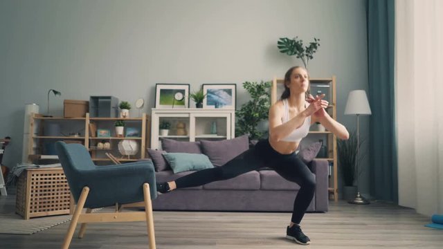 Pretty girl bodybuilder is exercising at home doing squats with armchair focused on exercise. Modern young people, healthy lifestyle and interior concept.
