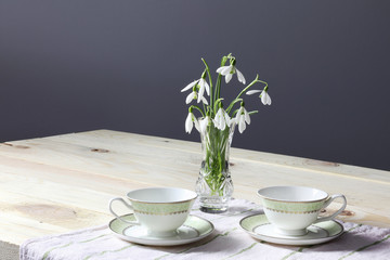 Snowdrops on the table. Spring flowers bouquet. Snowdrops background. Spring flowers on the wooden table. Holiday desk. Holiday flowers. Vase with spring flowers. Bowl with snowdrops. Two cups 