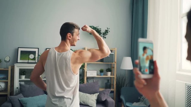 Cheerful guy handsome sportsman is posing for smartphone camera showing muscular body at home while young woman is holding device touching screen and talking.