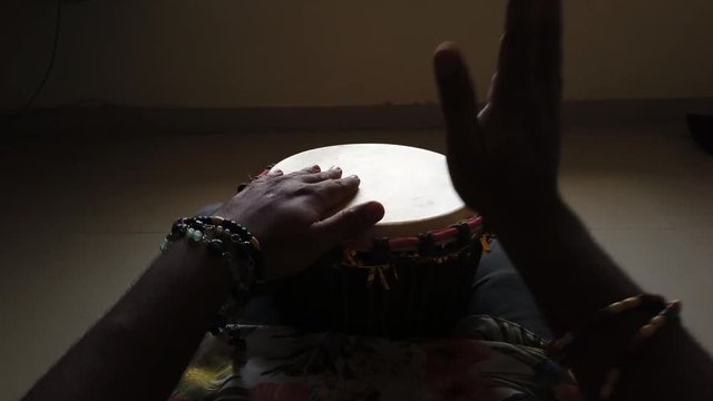 Medium wide looking down isolated shot of a man playing djembe drum alone in a dark room