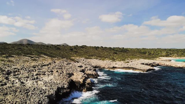 a drone shot flying backwards and showing waves splashing onto a rocky shore and spraying into the air near cala varques in majorca