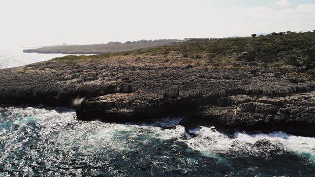 a forward flying drone shot flying towards a rocky shore with waves crashing onto the rocks and spraying into the air near cala varques in majorca