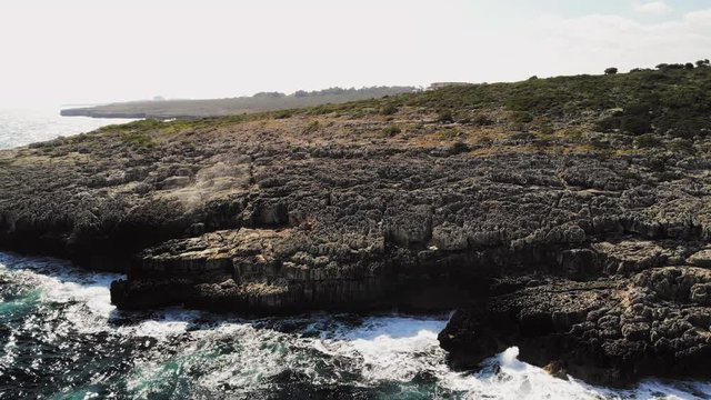 a drone shot starting close to a rocky shore with waves crashing and slowly flying to pan up and out near cala varques in majorca