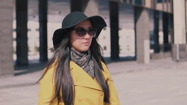stylish girl in a yellow coat with long dark hair in a hat walks down the street takes off his glasses and looks around