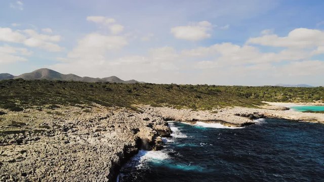 a drone shot flying backwards and showing waves splashing onto a rocky shore and spraying into the air near cala varques in majorca