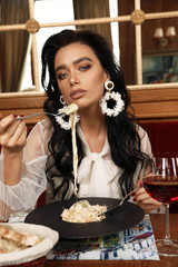 sensual woman with dark hair in elegant clothes and earrings sitting in restaurant, eating delicious italian pasta - 257611549
