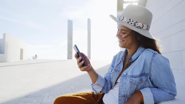 Portrait girl in hat with smartphone smiling and browsing or messaging while sitting on bench in park between modern building on sunny day