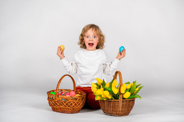Fototapeta na wymiar cute boy with wavy hair sits on a white background next to a basket with Easter eggs and holds two eggs in his hands