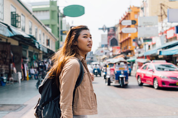 Young happy Asian female tourist backpacker walking and looking away on Khao San road in summer during trip to Bangkok, Thailand