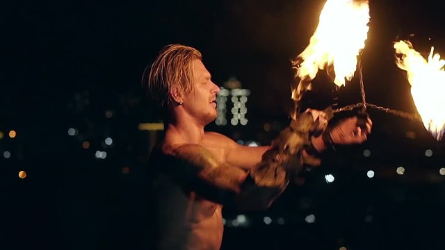 Young blond male spins two burning pois, ties them together. Slow motion shot. Close-up