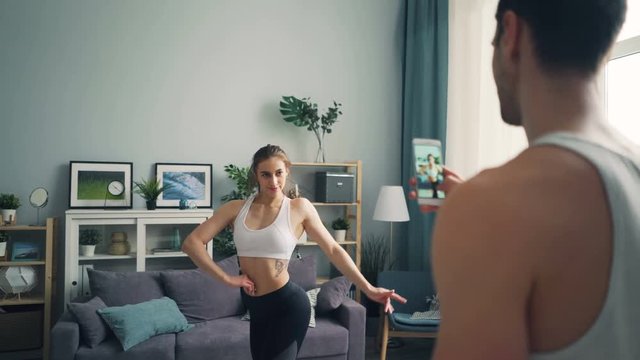 Young guy is recording video of pretty female bodybuilder posing for smartphone camera at home moving body and smiling. Smart phone screen is visible.