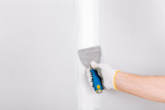 Man with putty knife shows how to hide the connection place between two pieces of dry walls using putty and construction tape
