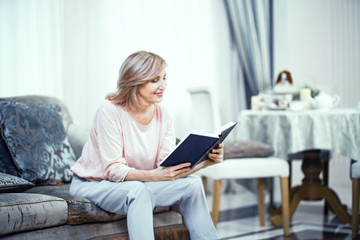 An Elderly Woman in Home Clothes is Sitting on the Sofa With a Book in Her Hands.