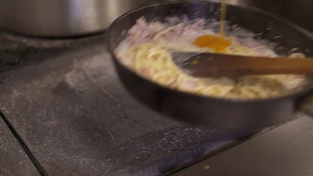 Handheld shot close up of spaghetti and bacon cooked in a cast-iron pan. Cook adding an egg to spaghetti frying on a pan on french cooking stove