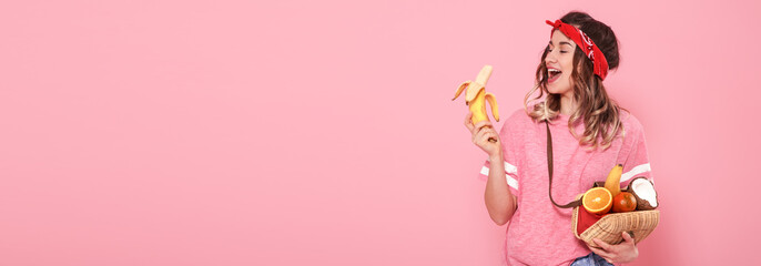 Beautiful young girl in pink t-shirt and glasses, holding sexy bananas and full bag of fruit on pink background