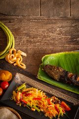 Papaya Salad (Som tum Thai) on square black plate placed on the wood table there are long bean, palm sugar, grilled fish, sticky rice and tomato placed around.