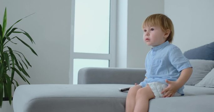 A 2 years old boy sits on a sofa and watches TV while sitting with the remote control in his hands. View cartoons and TV shows in childhood