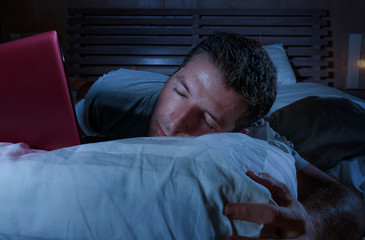 young tired and overworked man lying and networking on bed at home bedroom sleepy working with laptop computer late at night sleeping or falling asleep
