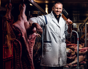 The butcher in a white work shirt holding ax and smiling while standing in a refrigerated warehouse in the midst of meat carcasses