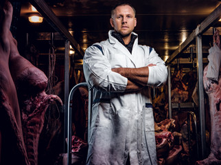 The butcher in workwear with his arms crossed in a refrigerated warehouse in the midst of meat carcasses