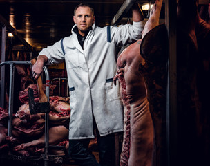 The butcher in a white work shirt holding ax while standing in a refrigerated warehouse in the midst of meat carcasses