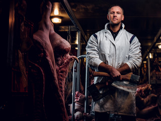 Fototapeta na wymiar The butcher in workwear posing with two axes in a refrigerated warehouse in the midst of meat carcasses