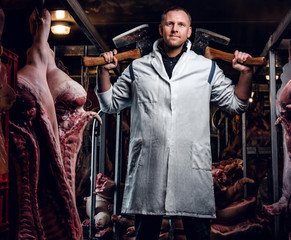 The butcher in workwear posing with two axes in a refrigerated warehouse in the midst of meat carcasses