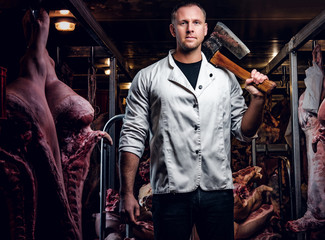 The butcher in a white work shirt holding ax while standing in a refrigerated warehouse in the midst of meat carcasses