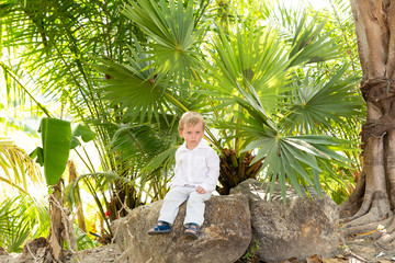 Naklejka na ściany i meble little blond boy in white clothes sitting on a stone under tropical trees