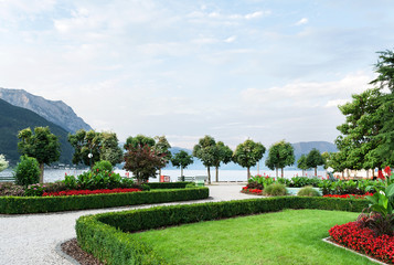 Obraz premium View of the mountains, the lake and the embankment of the city with trimmed trees, shrubs, lawn and flower beds.