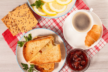 Toasted slices of bread with cheese and green parsley on white plate, cup of coffee, bowl with strawberry jam and spoon on napkin