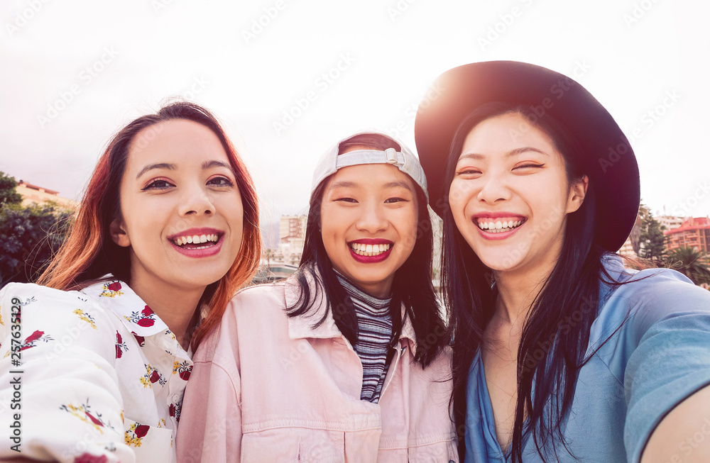 Wall mural Happy Asian girls taking selfie with mobile smartphone outdoor - Young social friends having fun taking self photos outside - People, millennial generation and youth lifestyle concept