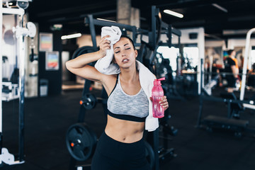 Young exhausted woman in fitness gym after successful workout wipes sweat from the forehead with white towel while holding water bottle in other arm. - obrazy, fototapety, plakaty