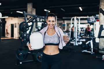 Fototapeta na wymiar Young attractive woman after successful workout posing and smiling in modern fitness gym while holding big white protein jar and glass of shake with drinking straw in other hand.