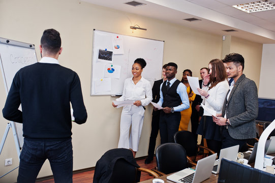 Mixed race business coach presenting report standing near whiteboard pointing on sales statistic shown on diagram and chart teach diverse company members gathered together in conference room.