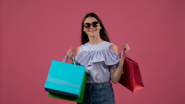 Gir in sunglasses l after shopping dancing with colorful packages . Pink background. Slow motion